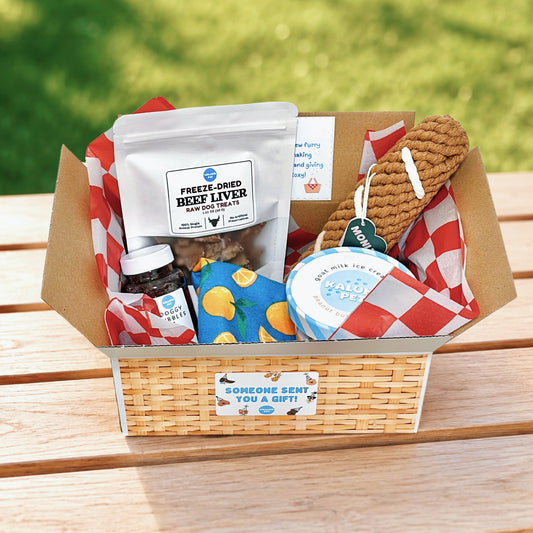 A curated pet gift basket displayed on a wooden outdoor bench with a blurred green lawn background. The basket contains a bag of freeze-dried beef liver, a jar of pet supplement, a blue and yellow patterned treat pouch, a round blue container, and a brown chew toy, all arranged in a woven wicker basket with red-and-white checkered paper lining.
