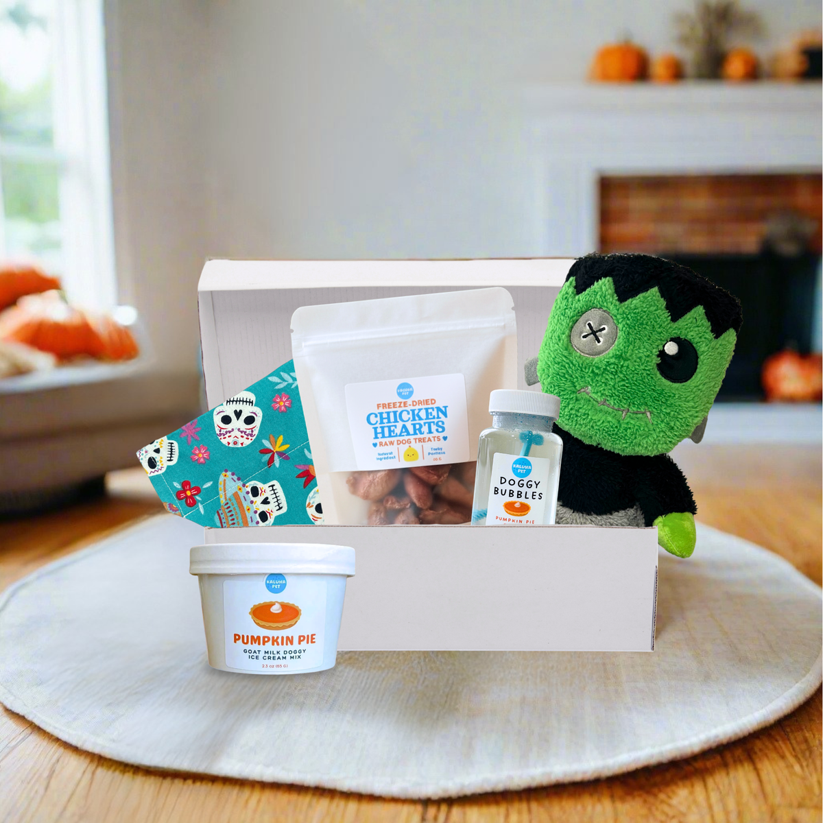 A curated Halloween gift box set displayed on a round white placemat over a wooden table, with a blurred living room background. The set includes a white box containing a green plush monster toy, a bag of Freeze-Dried Chicken Hearts, a small bottle of Dog Bubbles, a tub of Pumpkin Pie dog treats, and a colorful patterned bandana.