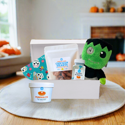 A curated Halloween gift box set displayed on a round white placemat over a wooden table, with a blurred living room background. The set includes a white box containing a green plush monster toy, a bag of Freeze-Dried Chicken Hearts, a small bottle of Dog Bubbles, a tub of Pumpkin Pie dog treats, and a colorful patterned bandana.